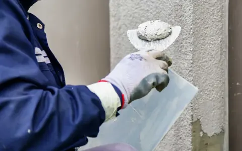 Pracownik w niebieskiej kurtce wykonuje tynkowanie elewacji, nakładając zaprawę kielnią na ścianę budynku.