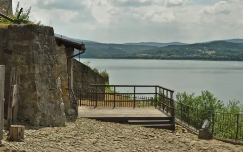 Kamienna wieża i drewniany taras z widokiem na spokojne jezioro dobczyckie. W oddali zalesione wzgórza.