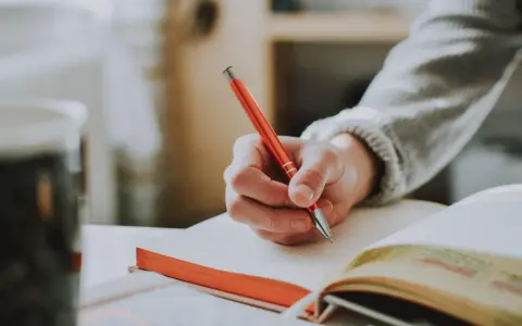 Main tenant une plume rouge, une main écrit dans un carnet. Un synonyme de participer pourrait être collaborer, comme dans cette scène d'étude.