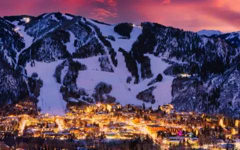 Glitzerndes Aspen, Colorado, im Abendrot. Schneebedeckte Berge mit Skipisten über einer Stadt, die in warmem Licht erstrahlt.