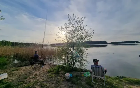 Dwóch wędkarzy siedzi nad jeziorem Wdzydze, czekając na branie ryb. Słońce zachodzi, malując niebo na złoto.