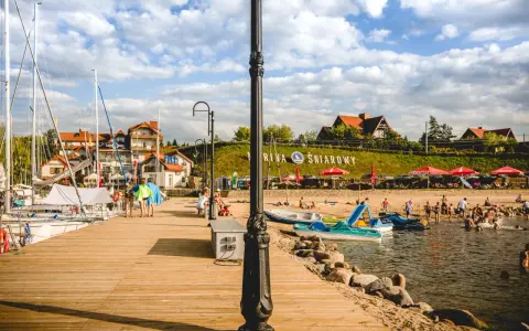 Słoneczna plaża nad jeziorem Śniardwy tętni życiem. Ludzie odpoczywają na piasku, kąpią się w wodzie, a obok cumują żaglówki.