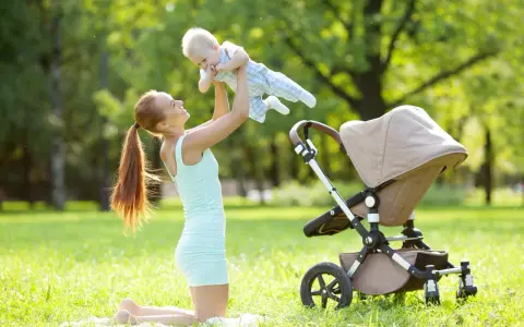 Uśmiechnięta mama podrzuca dziecko w powietrze na trawie obok wózka.