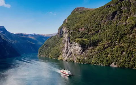 Ein Schiff fährt durch einen norwegischen Fjord im August. Das Wetter ist sonnig, die Berge sind grün und ein Wasserfall stürzt herab.