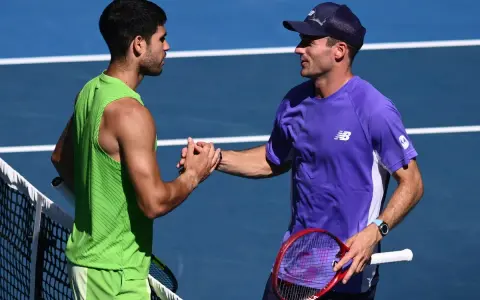 Dos tenistas, uno con camiseta verde y otro con morada, se dan la mano en la red. Tommy Paul, con gorra, sonríe tras el partido.