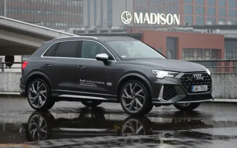 Szary Audi RS Q3 na mokrej nawierzchni, z odbiciem w kałuży. W tle budynek z napisem MADISON.