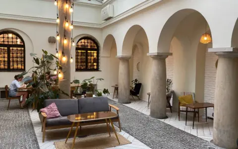 Przytulna kawiarnia patio z łukami i kolumnami. Goście siedzą przy stolikach, a obok stoi wygodna sofa. Widać rośliny i dekoracyjne lampki. Menu czeka na odkrycie.