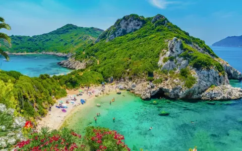 Malownicza zatoka z piaszczystą plażą, turkusową wodą, zielonymi wzgórzami i kwitnącymi kwiatami.
