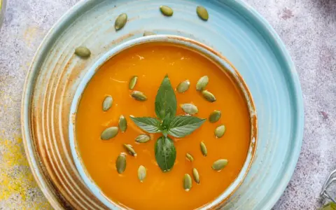 Zupa krem z dyni przepis Ewy Wachowicz. Aromatyczna, pomarańczowa zupa z pestkami dyni i listkiem mięty na niebieskim talerzu.