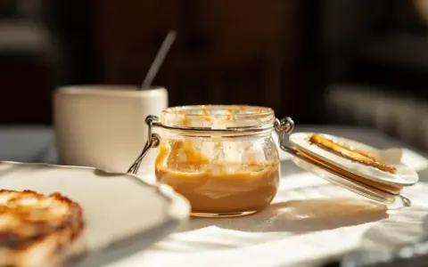 Glas mit cremiger Erdnussbutter, die gesund und lecker ist. Daneben ein Stück Toast.