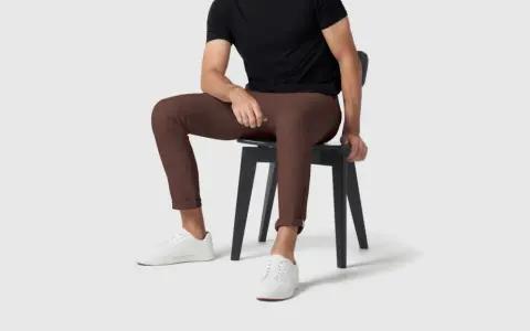 A man in a black shirt and brown pants sits on a black chair, wearing white sneakers.