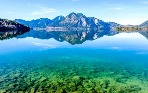 Kristallklarer Badesee in Bayern mit Felsen am Ufer und spiegelnden Bergen im Hintergrund.