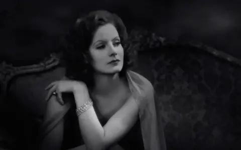 Elegancka Greta Garbo, z błyszczącą bransoletką, spogląda zamyślona z antycznej kanapy.