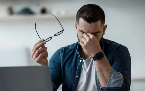 Homme fatigué, lunettes à la main, se frottant les yeux devant un ordinateur. Il est temps de penser à comment protéger ses yeux des écrans.