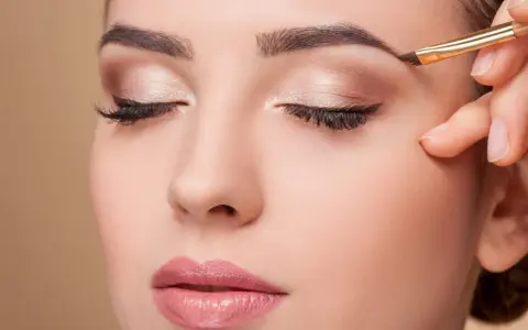 Une main applique un maquillage subtil pour parfaire la forme sourcil d'une femme aux yeux fermés.