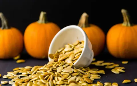Semi di zucca freschi versati da una ciotola, con zucche arancioni sullo sfondo. Perfetti per essere piantati.