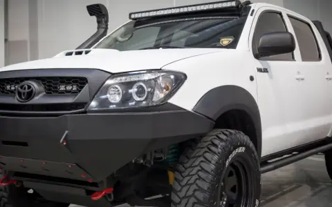 Biała Toyota Hilux z potężnym stalowym zderzakiem, wyciągarką i snorkel'em. Idealna do terenowych wypraw, gdzie przepisy nie ograniczają.