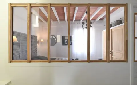 Une verrière en bois sépare deux pièces. On y voit un lit, des volets, une armoire et un lustre.