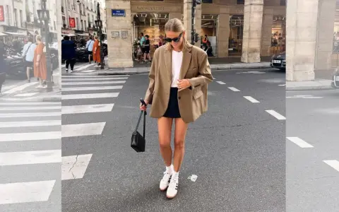 Mujer con look con blazer beige, falda negra y zapatillas blancas cruza un paso de peatones en París.