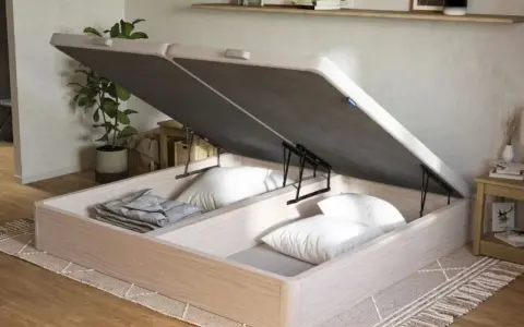 Un canapé de madera clara con la tapa abierta, revelando un gran espacio de almacenamiento para ropa de cama y almohadas.