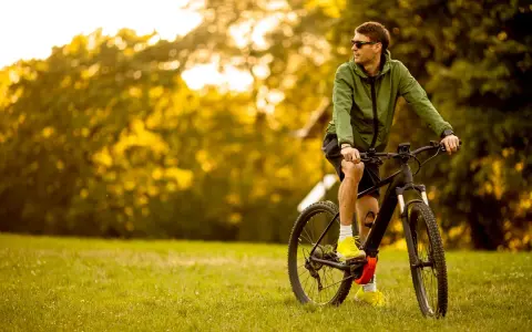Mężczyzna w zielonej kurtce i okularach przeciwsłonecznych odpoczywa na trawie obok swojego roweru elektrycznego ebike.