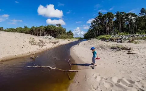 Dziecko w kasku spaceruje po piaszczystej plaży nad jeziorem Kopań. Atrakcje przyrodnicze i spokój.