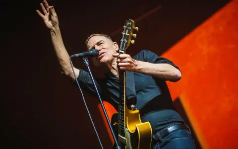 Bryan Adams na koncercie, w czarnej koszuli, z gitarą i mikrofonem, wznosi rękę w geście pozdrowienia.