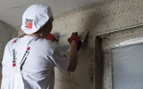 Pracownik w białej czapce i rękawiczkach gąbkuje tynk. Czas na gąbkowanie tynku zależy od jego rodzaju i warunków.