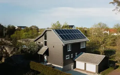 Ett modernt hus med mörk fasad och ett tak täckt av solceller. Bredvid huset finns ett garage med vita portar.