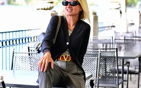 Donna con pantalone grigio elegante, abbinato a cardigan scuro e cintura con logo dorato. Un look chic per ogni occasione.
