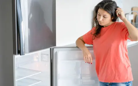 Une femme perplexe regarde son frigo vide, qui fait un bruit étrange. Elle se gratte la tête, se demandant ce qu'elle va manger.