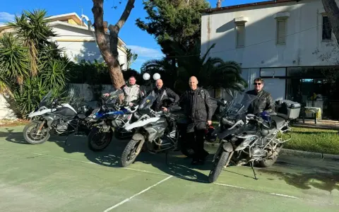 Czterech motocyklistów w pełnym rynsztunku gotowych na **prom na Sycylię**. Motocykle BMW GS zaparkowane na zielonym placu, w tle budynek i palmy.