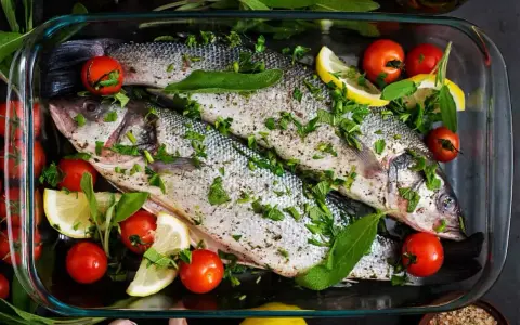 Dos lubinas enteras preparadas para ser horneadas, con tomates cherry, rodajas de limón y hierbas frescas.
