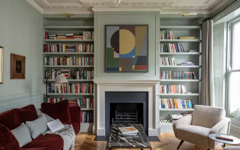 Un canapé au milieu du salon, entouré de bibliothèques remplies de livres, d'une cheminée et d'un fauteuil confortable.