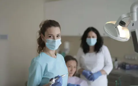Uśmiechnięta studentka stomatologii w masce i rękawiczkach trzyma wiertło dentystyczne. W tle inne osoby w gabinecie.