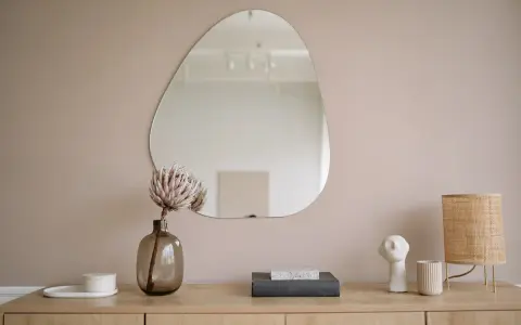 Un miroir vitrail aux formes organiques reflète une pièce épurée. Une fleur séchée dans un vase en verre fumé et une lampe en rotin complètent la scène.