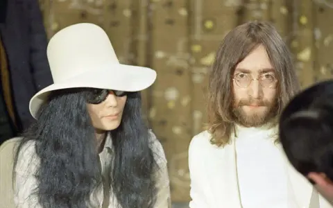 John Lennon och Yoko Ono, ikoniska konstnärer, poserar tillsammans. Yoko bär en stor vit hatt och solglasögon, medan John har sitt karaktäristiska långa hår och glasögon.
