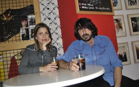 Pareja en un bar con decoración rockera. Ella, con camisa de cuadros, apoya la barbilla en la mano. Él, con camisa azul, sostiene un vaso. La furia trinidad de la música inspira el ambiente.