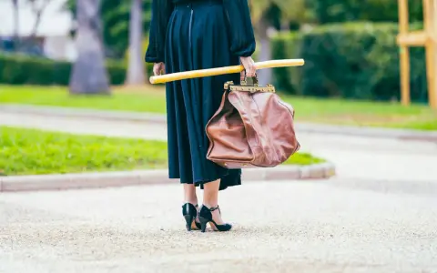 Elegancka kobieta w czarnej sukni i czarnych szpilkach, niosąca brązową torbę i żółty kij. Uosobienie francuskiej mody.