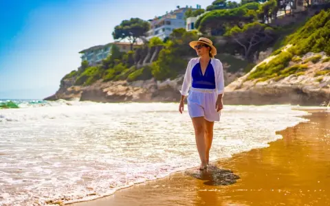Kobieta w słomkowym kapeluszu spaceruje po plaży. Idealne miejsce na tanie loty na majówkę, by poczuć słońce i fale.