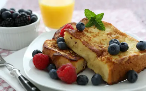Tostadas francesas doradas, decoradas con arándanos y frambuesas, junto a un vaso de jugo de naranja.