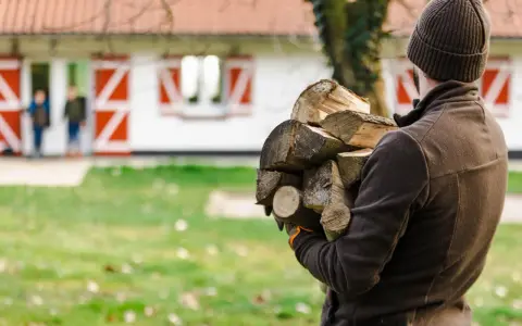 Chcesz wiedzieć, jak zostać leśniczym? Ten mężczyzna z drewnem na podpałkę, w tle dom z czerwonymi okiennicami i dzieci, pokazuje, że praca w lesie to też praktyczne zadania.