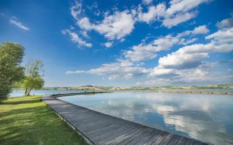 Passeggiata in legno sul **lago di Chiusi**, con cielo azzurro e nuvole bianche che si riflettono sull'acqua calma.