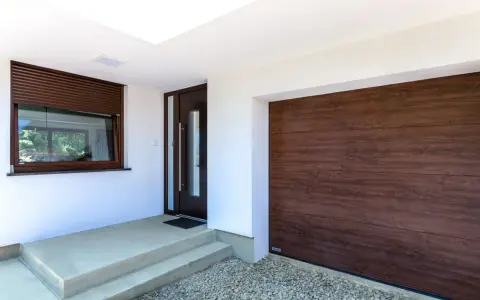 Ingresso con porta scura e avvolgibili marroni, accanto a un'ampia porta garage in legno scuro.