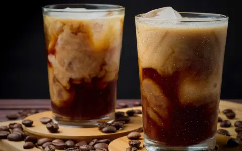 Två glas med iskaffe, perfekt för ett iskaffe recept. Krämig mjölk virvlar i mörkt kaffe med isbitar. Kaffebönor sprider sig runt glasen.
