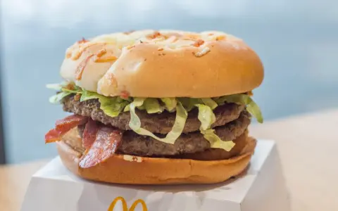 Duży burger z dwoma kotletami, bekonem i sałatą na białym opakowaniu.