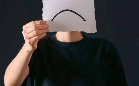 Osoba w czarnej bluzie trzyma kartkę z narysowaną smutną buźką. Czy to anhedonia, co to za uczucie?