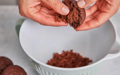 Hände zerbröseln einen Schokoladenkuchen für vegane Desserts. Ein Teil landet in einer weißen Schüssel.