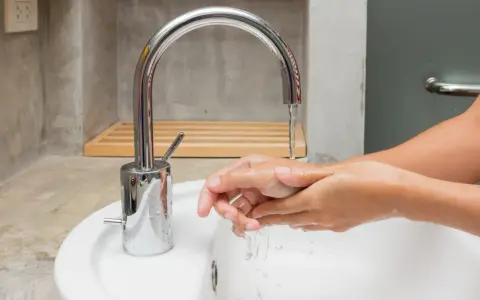 Mains d'une personne se lavant sous un robinet moderne en inox. L'eau coule dans un lavabo blanc.