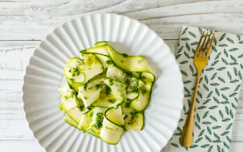 Delicato carpaccio di zucchine condito con erbe fresche, servito su un piatto bianco con forchetta dorata.